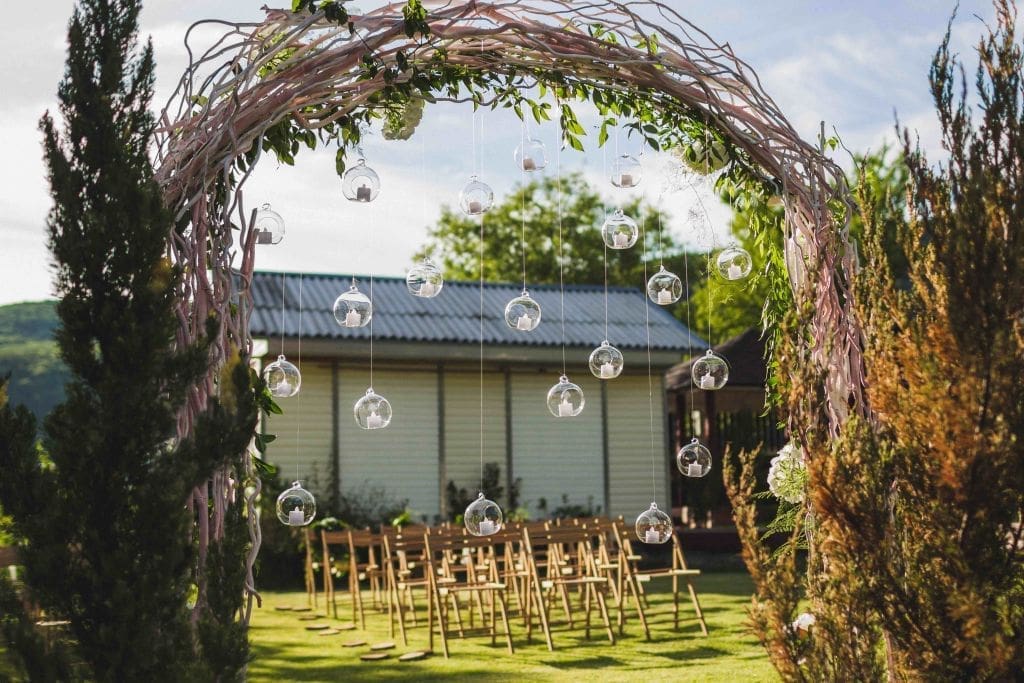 Evening wedding ceremony in garden, arch with white branches, brown wooden chairs and a lot of lights hanging in glass spheres, wedding catering options, Sydney.