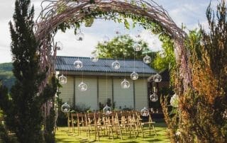 Evening wedding ceremony in garden, arch with white branches, brown wooden chairs and a lot of lights hanging in glass spheres, wedding catering options, Sydney.